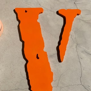 Op de afbeelding: Een grote, oranje, gestileerde letter V gemaakt van hout. De letter is geverfd in een helder oranje en heeft een ruw, getextureerd oppervlak.
