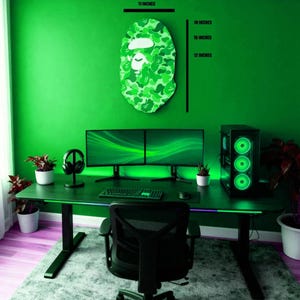 May include: A green-themed gaming setup featuring a camouflage-patterned face art piece, measuring 45.7 cm in height and 27.9 cm in width. The room includes a black desk with dual monitors, a computer tower, and a black gaming chair. The wall is painted green.