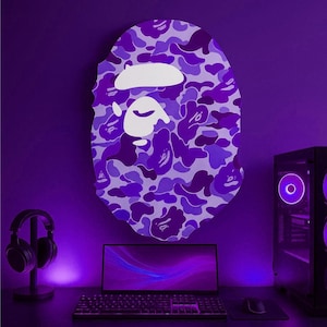 Puede incluir: Decoraci&oacute;n de pared con estampado de camuflaje morado y blanco en forma de rostro estilizado. La obra de arte mural est&aacute; montada sobre un escritorio con un port&aacute;til, teclado, rat&oacute;n y torre de ordenador. Un par de auriculares est&aacute; a la izquierda.