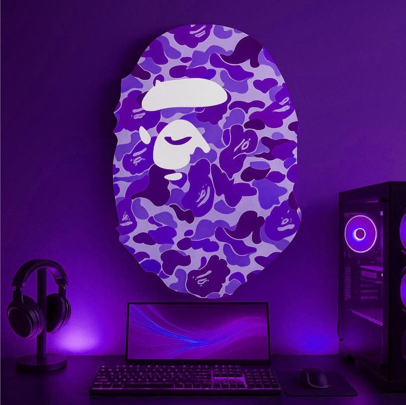Puede incluir: Decoraci&oacute;n de pared con estampado de camuflaje morado y blanco en forma de rostro estilizado. La obra de arte mural est&aacute; montada sobre un escritorio con un port&aacute;til, teclado, rat&oacute;n y torre de ordenador. Un par de auriculares est&aacute; a la izquierda.