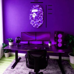 Puede incluir: Una configuraci&oacute;n de juego de color p&uacute;rpura con un escritorio, monitores duales, una torre de computadora y una silla negra. Una decoraci&oacute;n de pared con una cara de camuflaje p&uacute;rpura, 46 cm de alto y 28 cm de ancho. La habitaci&oacute;n est&aacute; iluminada con luces p&uacute;rpuras.