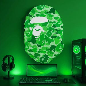 May include: Green camouflage-patterned wall art in the shape of a head, above a desk with a laptop, keyboard, and mouse. A computer tower and headphones are also visible. The room is illuminated with green lighting.