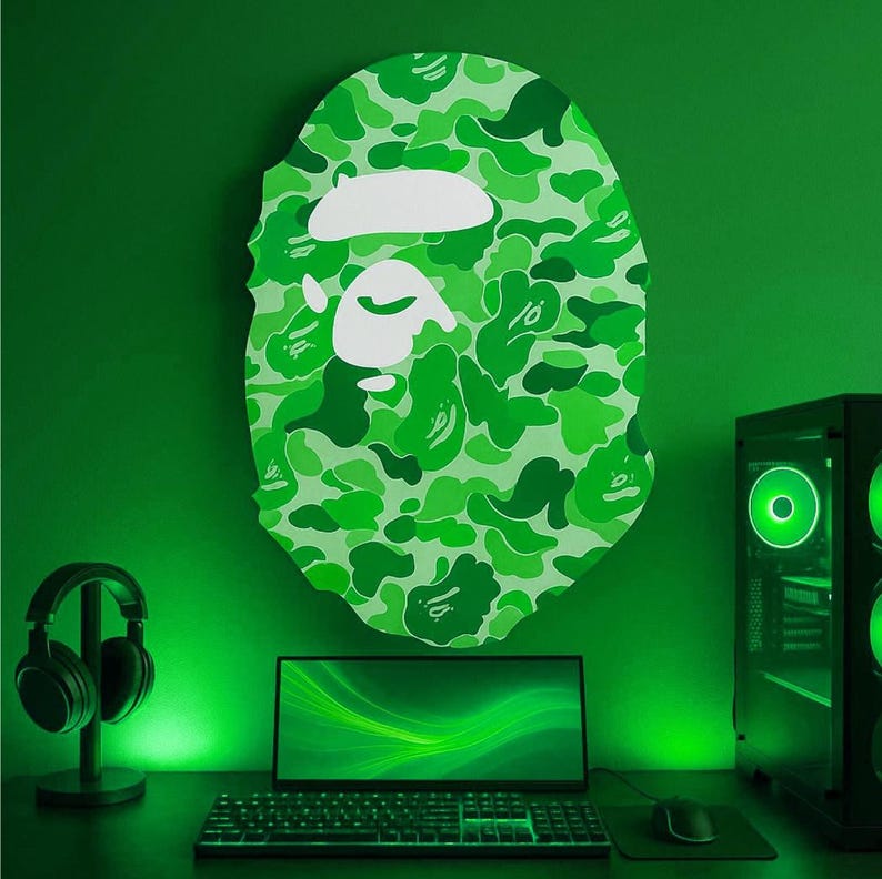 May include: Green camouflage-patterned wall art in the shape of a head, above a desk with a laptop, keyboard, and mouse. A computer tower and headphones are also visible. The room is illuminated with green lighting.