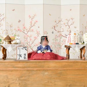 May include: A baby wearing a traditional Korean outfit sits on a red cushion in front of a white screen with floral designs. The baby is wearing a blue and white top with a black hat. There are two white vases with white flowers on either side of the baby. There is a wooden table in front of the baby with a small wooden box with Korean writing on it.