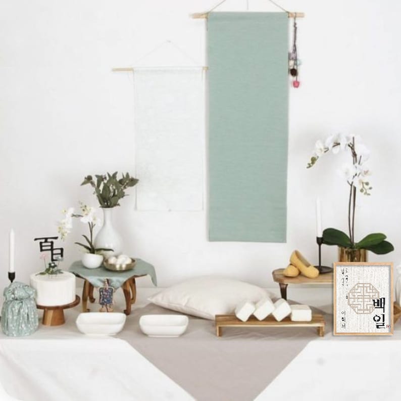 May include: A traditional Korean Dol ceremony table setting with a white cake, white rice cakes, a white vase with green leaves and white flowers, and a framed calligraphy piece with the Korean word "Dol" in black ink.