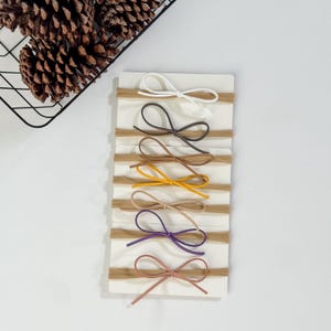 May include: A collection of seven baby headbands with small bows in various colors, including white, brown, yellow, tan, purple, and pink. The headbands are arranged on a white rectangular card, with a basket of pine cones in the upper left corner.
