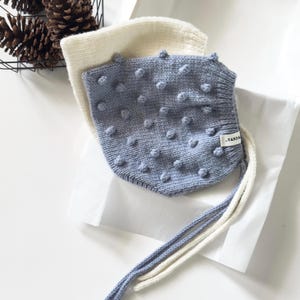 May include: Two knitted baby bonnets. One is light blue with textured bobbles, the other is off-white. Both have long ties. The light blue bonnet has a small tag that reads "-YARDA".