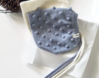 May include: A blue knitted bonnet featuring a textured bobble pattern across the surface and a small white fabric tag with the text VARDA. The bonnet includes long, cream-colored knitted ties and is layered over a plain cream-colored knitted bonnet.