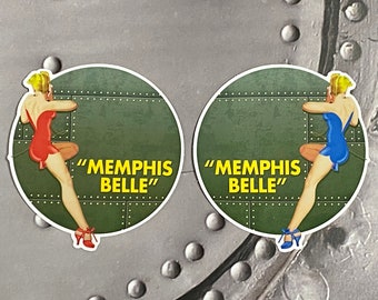 May include: Two round stickers with a green background and a pin-up girl design. The stickers feature the text "Memphis Belle" and a woman in a red dress and a woman in a blue dress.