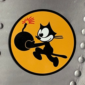 May include: A circular sticker featuring the cartoon character Felix the Cat holding a lit bomb. The image has a black outline, a yellow background, and is set against a silver background with rivets.