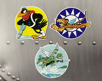 May include: Three colorful round stickers featuring cartoon animal mascots. The first sticker features a black bird wearing a red scarf and goggles, flying with a bomb. The second sticker features a tiger with wings flying in front of a blue circle with white stars. The third sticker features two green turtles with wings, flying with a sword.