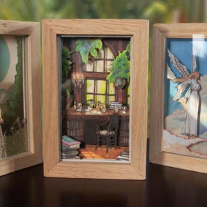 Può includere: Tre opere d'arte incorniciate con dettagliati ritagli di carta. Le cornici sono in legno chiaro. L'arte raffigura scene di angeli, una donna in un giardino e una stanza con scrivania e sedia. Gli sfondi sono dipinti con colori tenui.
