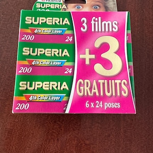 Puede incluir: Un paquete de película en color Fujifilm Superia 200. El paquete es verde y rosa con el texto "3 films + 3 gratuits" y "6 x 24 poses". La película es 4th Color Layer.