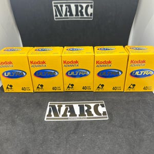 Op de afbeelding: Vijf gele en zwarte filmcassettes met het label "Kodak Advantix Ultra" met "APS" en "40 EXP FILM" erop gedrukt. De cassettes zijn in een rij op een zwart oppervlak gerangschikt.