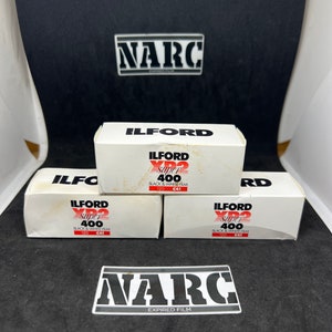 Könnte beinhalten: Drei weiße Schachteln mit Ilford XP2 Super 400 Schwarzweißfilm, mit roter und schwarzer Schrift. Die Schachteln sind mit "120 C41" beschriftet. Das Bild enthält auch zwei "NARC Expired Film" Aufkleber.