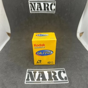 Op de afbeelding: Een gele Kodak Advantix Ultra 200 filmdoos met blauwe en witte tekst. De doos heeft de tekst "ULTRA" in een blauwe ovaal en "40 EXP FILM" onderaan. De achtergrond is donkergrijs met de tekst "NARC" boven en onder.