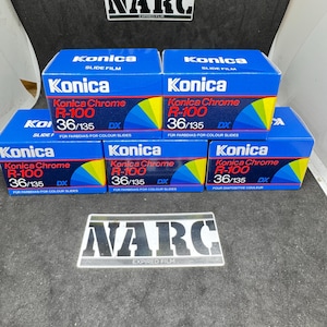 Op de afbeelding: Een verzameling Konica Chrome R-100 36/135 DX diafilmdozen. De blauwe dozen hebben gele en groene accenten en het Konica-logo. Er is ook een NARC-sticker voor verlopen film te zien.