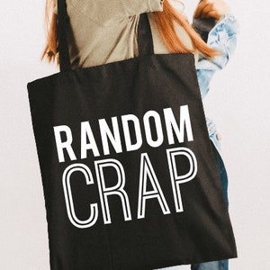 May include: Black tote bag with the words "RANDOM CRAP" in large white block letters. The bag has two black handles and is held by a person. The background is a plain white wall.