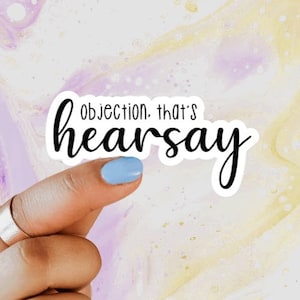 May include: A white sticker with the text "objection, that's hearsay" in black script and sans-serif font. The sticker is held by a finger with a light blue nail polish and a silver ring, against a colorful background.