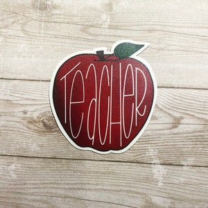 May include: Red apple sticker with the word "Teacher" written in white cursive lettering.