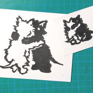 Puede incluir: Impresión en linóleo en blanco y negro de dos perros, uno de pie y otro acostado. La impresión está sobre un fondo blanco.