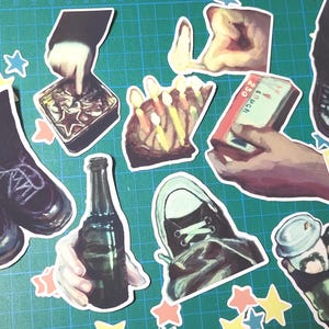 May include: A collection of colourful stickers featuring various images, including a pair of black boots, a bottle of beer, a hand holding a matchbox, a hand holding a can of soda, a hand holding a birthday cake with candles, a hand holding a shoe, and a hand holding a cup of coffee. The stickers are on a green background with stars.