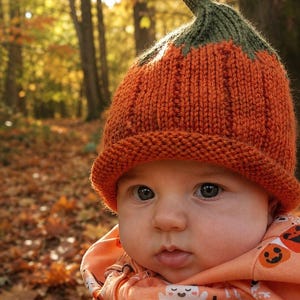 May include: A knitted orange hat with a green stem-like top, featuring a ribbed texture and a rolled brim. The hat is worn with a light orange hooded garment decorated with a repeating pattern of small white ghosts and orange jack-o'-lantern faces.
