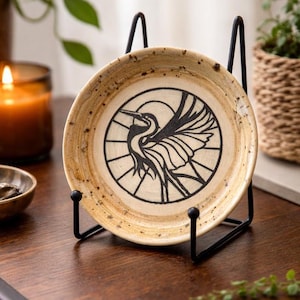 May include: A small, round ceramic plate with a black heron design, displayed on a black metal stand. The plate has a speckled, tan and brown surface. The heron is depicted with its wings spread, set against a circular background.