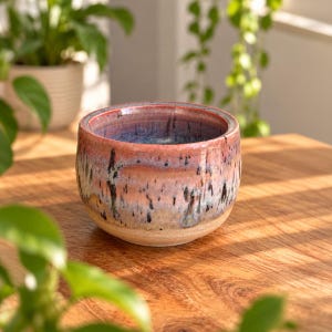 May include: A round ceramic bowl featuring a reactive glaze with blended hues of pink, peach, and blue. The exterior surface displays vertical dark streaks and drips, while the interior reveals a deep blue tone. The base is unglazed and light tan.