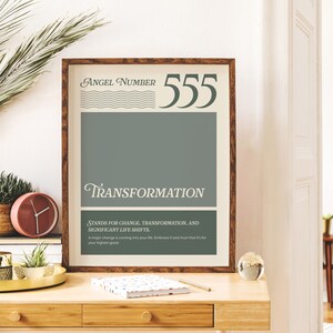 May include: A framed print with a beige background and brown frame. The print features the text "Angel Number 555" in a green font. Below the number, the word "Transformation" is written in a green cursive font. The text "Stands for change, transformation, and significant life shifts." is written in a smaller font below the word "Transformation". The text "A major change is coming into your life. Embrace it and trust that it's for your highest good." is written in a smaller font below the previous text.