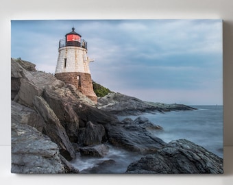 May include: A white lighthouse with a red roof stands on a rocky coastline. The water is a hazy blue and the sky is a light blue with gray clouds.