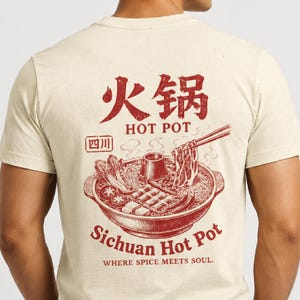 Pode incluir: Uma t-shirt de cor creme com um gráfico vermelho nas costas. O design mostra um hot pot de Sichuan com vegetais e carne, acompanhado pelo texto "火锅", "HOT POT", "四川", "Sichuan Hot Pot" e "WHERE SPICE MEETS SOUL."