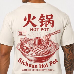 Pode incluir: Uma t-shirt cor de creme com um gráfico vermelho nas costas. O design mostra um hot pot de Sichuan com ingredientes, pauzinhos e o texto "火锅", "HOT POT", "四川", "Sichuan Hot Pot" e "WHERE SPICE MEETS SOUL" num estilo desgastado.