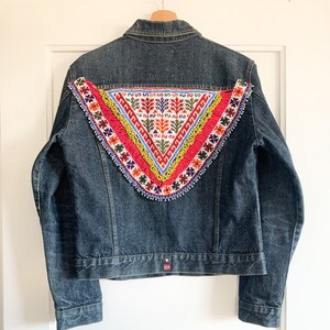 Könnte beinhalten: Eine dunkelblaue Jeansjacke mit einem bunten, gestickten Dreieck auf der Rückseite. Die Stickerei zeigt geometrische Muster in Rot, Gelb, Blau, Grün und Weiß.