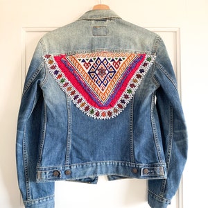 May include: A blue denim jacket with a colourful embroidered patch on the back. The patch features a geometric design with pink, orange, white and blue beads.