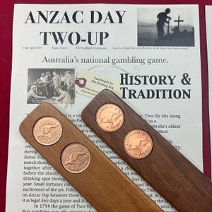 May include: Two wooden paddles with embedded Australian penny coins are displayed on a newspaper. The newspaper's headline reads "ANZAC DAY TWO-UP" with text about Australia's national gambling game. The paddles are a dark brown colour.