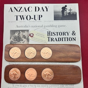 Może przedstawiać: Drewniana deska z trzema australijskimi monetami o wartości jednego pensa z 1965, 1961 i 1951 roku. Deska jest opisana jako "ANZAC DAY TWO-UP" i "Narodowa gra hazardowa Australii." Deska została wykonana ręcznie przez "the droving dog".