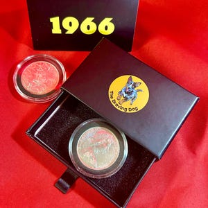 May include: A silver coin in a clear case is displayed inside a black box with a yellow sticker featuring a blue heeler dog. The box is open, revealing a coin. A separate coin in a clear case and a box with "1966" are also visible.