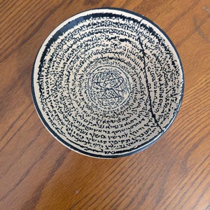 May include: A round ceramic bowl with a black rim and a beige interior. The bowl is decorated with black text in a circular pattern. A crack runs from the rim towards the center. The bowl is on a wooden surface.