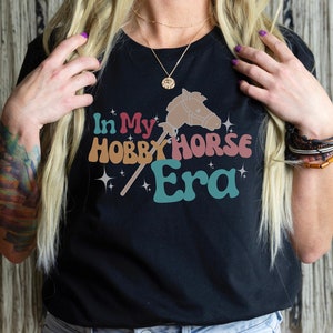 May include: A black t-shirt with the text "In My Hobby Horse Era" in colorful retro font. The text is accompanied by a graphic of a hobby horse and stars.