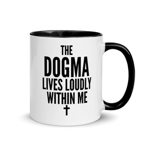 May include: White ceramic coffee mug with a black rim and handle. The mug has black text that reads "The Dogma Lives Loudly Within Me" with a black cross underneath.