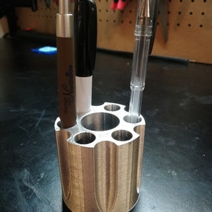 May include: A silver-colored pen holder with multiple circular compartments. The holder contains a brown pen with the text "Women of Northside", a black marker, and a clear pen. The holder is cylindrical with a unique, ridged design.