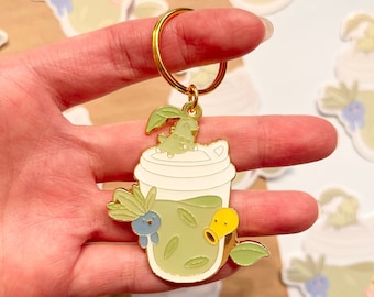 May include: Gold keychain featuring a green tea-themed enamel pin. The pin depicts a green tea drink with a green plant growing out of the top, a yellow and blue character inside the drink, and green leaves around the cup.