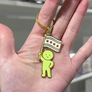 May include: A green keychain featuring a small figure holding a flag with the Chicago city flag design. The keychain has a gold-colored ring. The figure is a light green color.