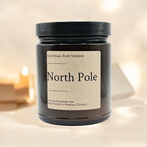 May include: A brown glass candle jar with a black lid. The label reads "Spiritual Kids Candles" and "North Pole". The label also states "Natural Soy Candle Box" and "Hand Poured in Bradley, Illinois".
