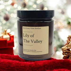 Puede incluir: Un tarro de vela de vidrio marr&oacute;n con una tapa negra. La etiqueta dice "Spiritual Kids Candles Lily of The Valley Natural Soy Candle Box Hand Poured in Bradley, Illinois".