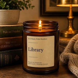 May include: A lit soy candle in an amber glass jar with a cream-coloured label reading Spiritual Kids Candles, Library, and Natural Soy Candle 8oz, Hand Poured in Bradley, Illinois. The candle sits beside a stack of three vintage books.