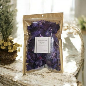 May include: A clear plastic bag filled with dried purple flower petals. The bag is labeled "Spiritual Kids Lavender + Cedar Potpourri".