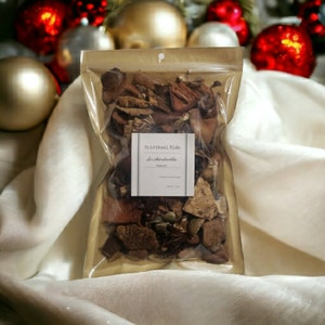 May include: A clear plastic bag filled with dried botanicals, with a label that reads "Spiritual Kids" and "Snickerdoodle". The bag is sealed at the top and sits on a white, textured surface. Christmas ornaments are in the background.