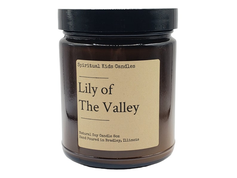 Puede incluir: Un tarro de vela de vidrio marr&oacute;n con una tapa negra. La etiqueta es marr&oacute;n con texto blanco que dice "Spiritual Kids Candles", "Lily of" y "The Valley". La etiqueta tambi&eacute;n incluye el texto "Natural Soy Candle 6oz" y "Hand Poured in Bradley, Illinois".